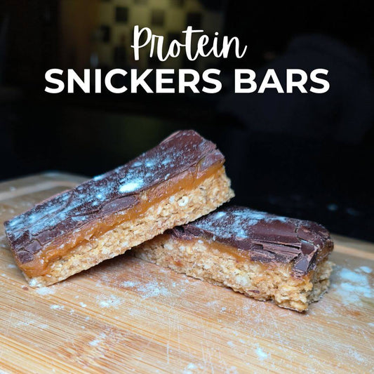 A protein bar resembling a Snickers bar, cut in half to show the layers, placed on a wooden surface with the text 'Protein SNICKERS Bars' above it.